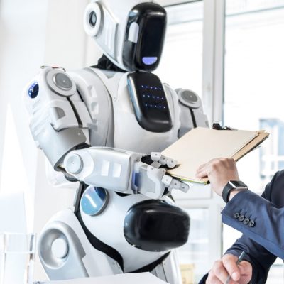 Futuristic helper. Low angle of serious middle-aged businessman sitting at table in office while big robot is passing him important documents